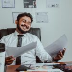 Un homme souriant tient des documents devant un ordinateur