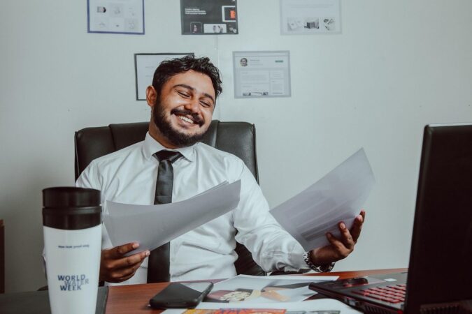 Un homme souriant tient des documents devant un ordinateur