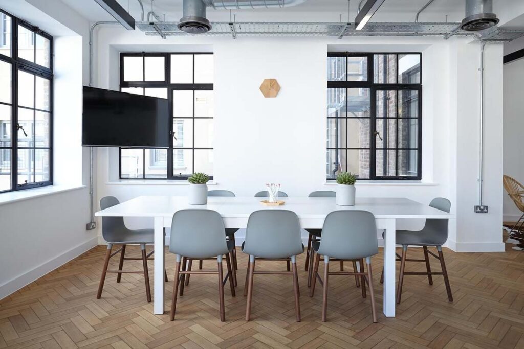 Une salle lumineuse avec table blanche, chaises, fenêtres et téléviseur mural