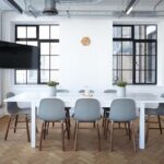 Une salle lumineuse avec table blanche, chaises, fenêtres et téléviseur mural