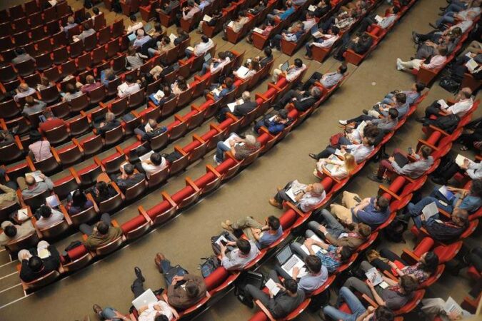 Salle de conférence avec personnes assises sur des sièges rouges, certaines utilisent des ordinateurs portables