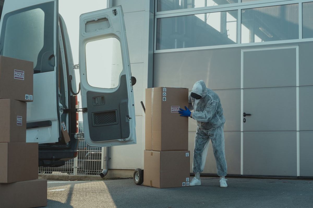 Individu en blouse blanche manipule des boîtes en carton près d'un van devant un édifice Individu en blouse blanche manipule des boîtes en carton près d'un van devant un édifice